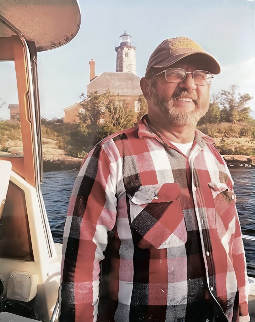 A man wearing glasses, a cap, and a red plaid shirt stands on a boat near the shore with a stone lighthouse and trees in the background under a clear sky.