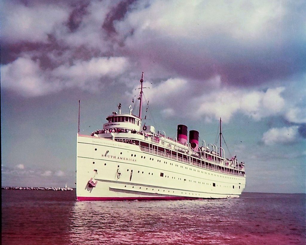 A large white passenger ship labeled "South American" sails on calm water under a partly cloudy sky, with two red and black smokestacks visible on the deck.