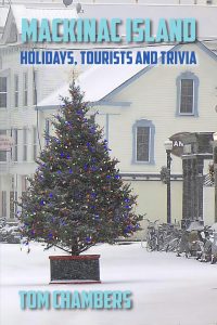 A snowy street scene on Mackinac Island features a large, decorated Christmas tree in the foreground and historic buildings in the background. The book title and author’s name are overlaid in bold blue text.