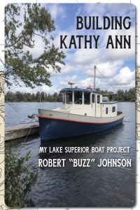 A small blue and white boat named Kathy Ann is docked on a lake under a partly cloudy sky. Trees line the shore in the background. Text on the image reads: "Building Kathy Ann, My Lake Superior Boat Project, Robert 'Buzz' Johnson.