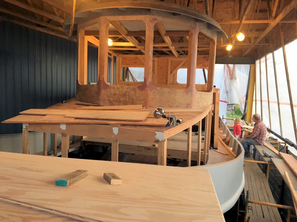 A wooden boat under construction in a covered workshop, with tools and planks on the deck and an unfinished cabin structure. A person sits at a table in the background near the right side of the image.
