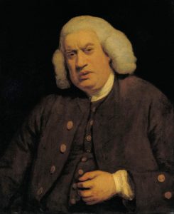 A man with white, shoulder-length hair wears a dark brown coat over a waistcoat and shirt. He has a serious expression and is seated against a dark background.