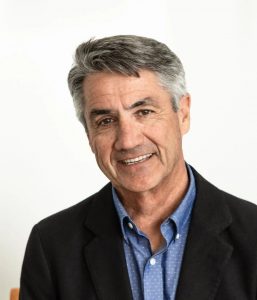 A middle-aged man with gray hair is wearing a black blazer over a blue shirt with a subtle pattern. He is smiling and sitting in front of a plain white background.
