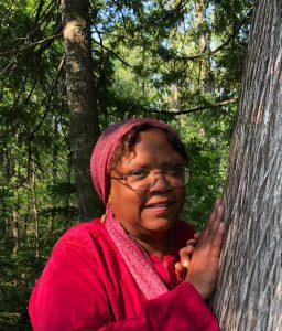 A woman wearing glasses, a red headscarf, and a red shirt stands outdoors, leaning against a tree with both hands. Green trees and sunlight are visible in the background.