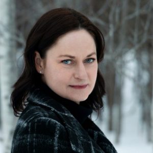 A woman with fair skin and dark hair wearing a dark coat stands outdoors in a snowy, wooded area, looking at the camera with a neutral expression.