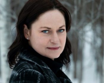A woman with fair skin and dark hair wearing a dark coat stands outdoors in a snowy, wooded area, looking at the camera with a neutral expression.