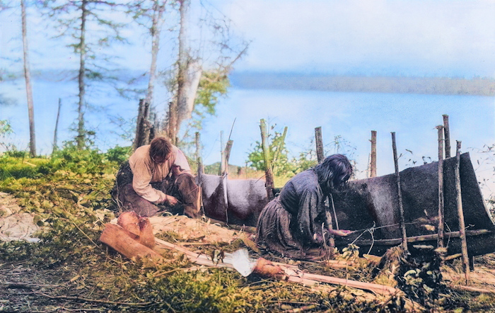 Two people in worn clothing work on constructing a canoe-like structure near a lakeshore, surrounded by trees and wooden poles, with calm water visible in the background.