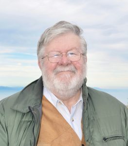 An older man with gray hair, glasses, and a white beard is wearing a green jacket over a tan vest and white shirt, standing outside with a cloudy sky in the background.