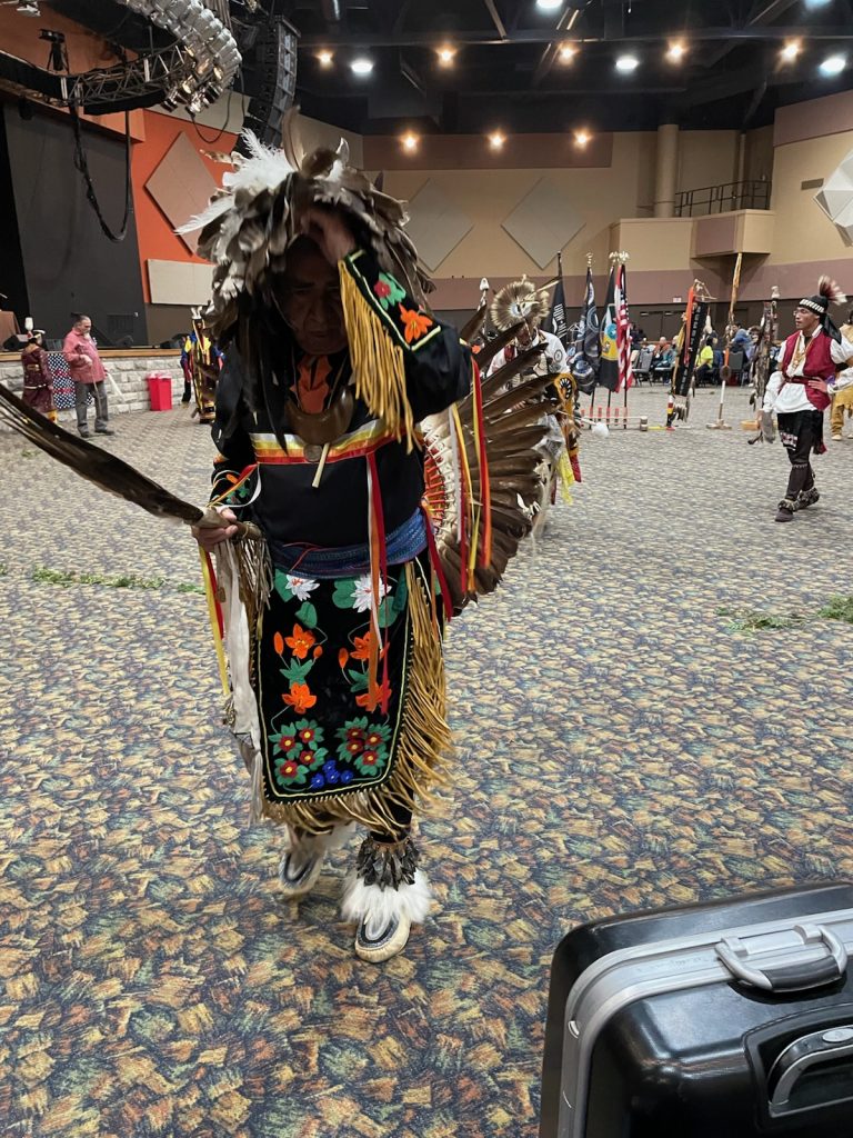 A person in traditional Native American regalia with feathered accessories dances indoors on a patterned carpet. Other similarly dressed people and flags are visible in the background.