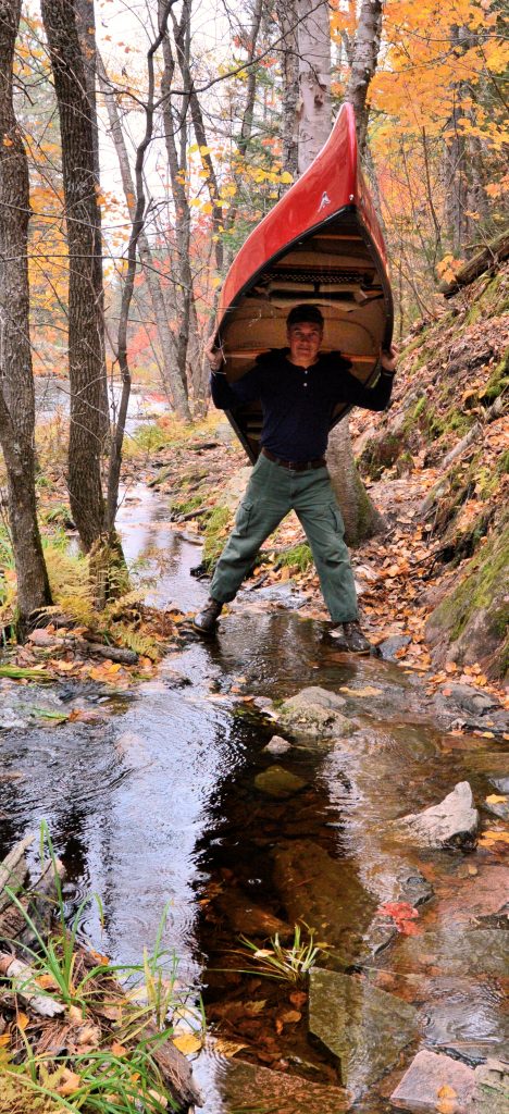 A person wearing a dark shirt and green pants stands on rocks in a creek, carrying a red canoe overhead. Trees with autumn leaves and a forested hillside surround the scene.