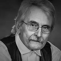 An older man with light hair, glasses, and a mustache looks at the camera. He is wearing a light-colored shirt and a dark vest. The image is in black and white.