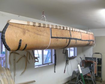 A birchbark canoe is suspended upside down from the ceiling of a garage or workshop, held by ropes. Tools, lumber, and equipment are visible on the walls and floor.
