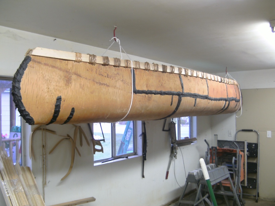 A birchbark canoe is suspended upside down from the ceiling of a garage or workshop, held by ropes. Tools, lumber, and equipment are visible on the walls and floor.