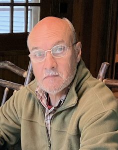 An older man with a bald head, gray beard, and glasses sits indoors wearing a green fleece jacket over a plaid shirt. Wooden chairs and a window are visible in the background.