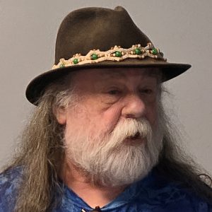 An older man with a long white beard and hair wears a brown hat decorated with beads and a blue shirt. He is facing slightly left against a plain background.