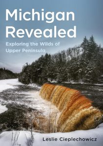 Book cover for "Michigan Revealed: Exploring the Wilds of Upper Peninsula" by Leslie Ciepłechowicz, featuring a river waterfall surrounded by snow-covered trees under a cloudy sky.