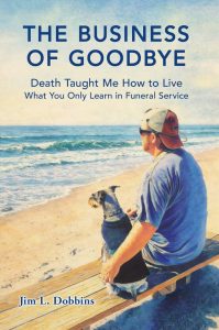 A man in a blue shirt and cap sits on a bench at the beach with a small dog, looking at the ocean. The book title "The Business of Goodbye" and author Jim L. Dobbins are visible.